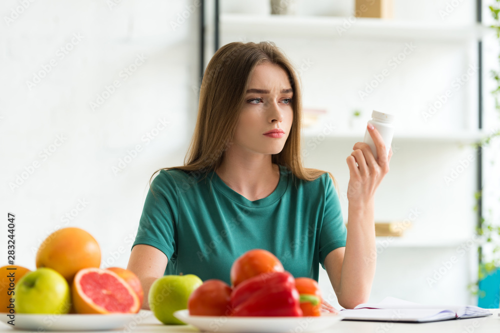 sad woman sitting at table with fruits and vegetables and holding pills