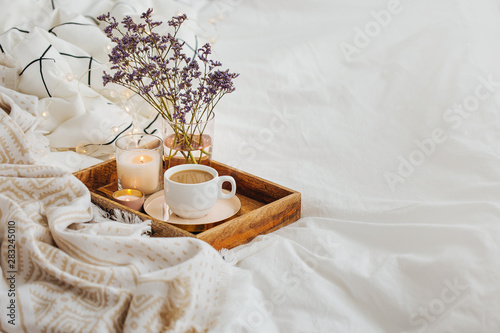 Wooden tray of coffee and candles with flowers on bed. White bedding sheets with striped blanket and pillow. Breakfast in bed. Hygge concept. photo