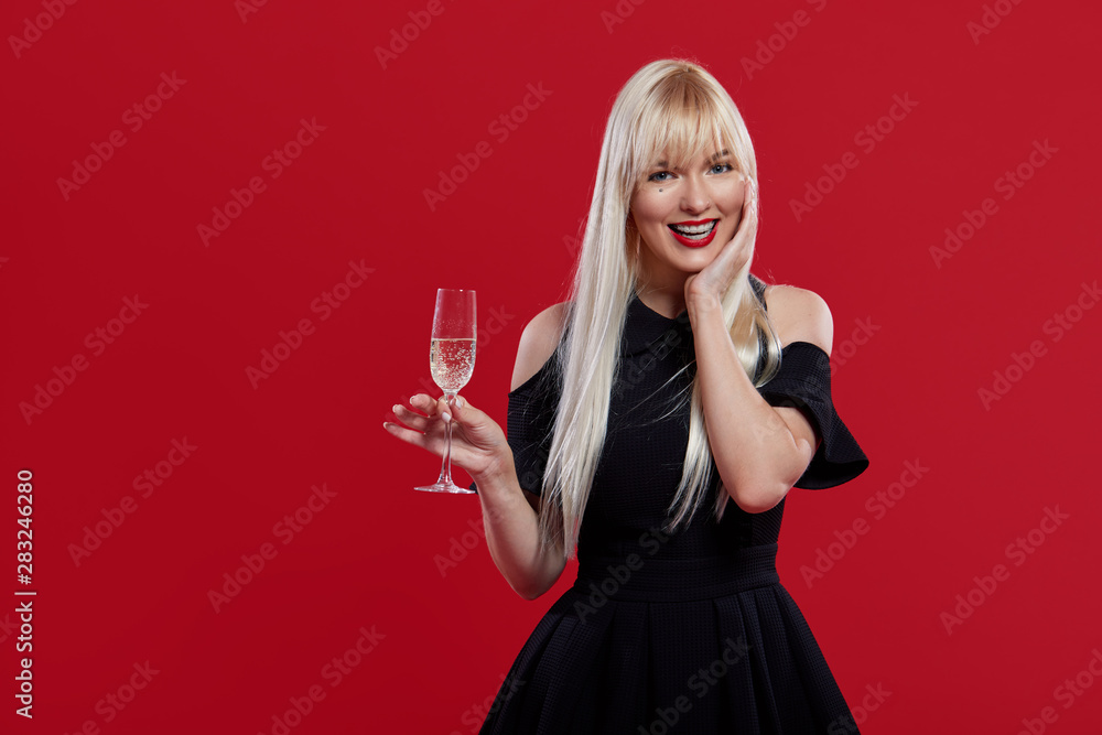 Party time! Portrait of young celebrating woman in black dress. Charming, lovely blond woman with champagne in hands isolated on red background. Woman in braces and with snow-white smile Party time! Portrait of young celebrating woman in black dress. Charming, lovely blond woman with champagne in hands isolated on red background. Woman in braces and with snow-white smile