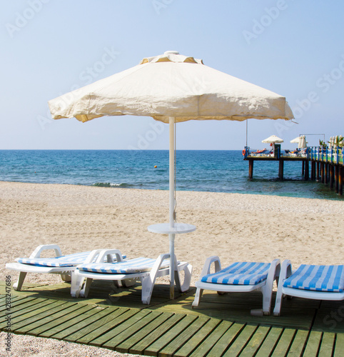 Sun loungers under the beach umbrella on the sandy beach. White deck chairs with mattresses under the awning. Sea holidays in southern countries
