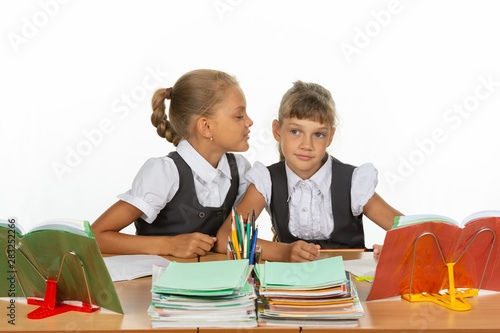 Girls are sitting at a school desk, one whispers to the other something in the ear photo