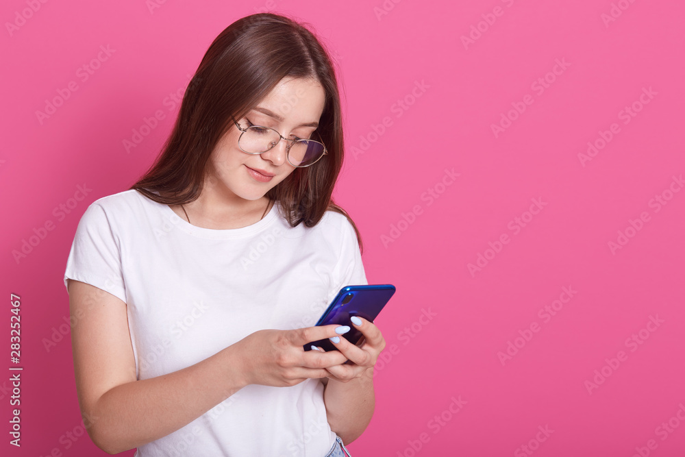 Indoor shot of young good looking European woman isolated on rosy background holding blue phone in hands and looking to its screen, reading message from her boyfriend. Modern technology concept. Indoor shot of young good looking European woman isolated on rosy background holding blue phone in hands and looking to its screen, reading message from her boyfriend. Modern technology concept.