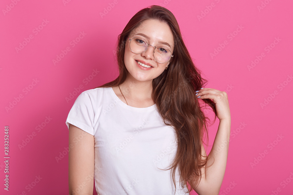 Smiling woman in eyeglasses posing in studio against pink wall looking directly at camera, tilts head and touches her skein of hear, has pleasant appearance, being in good mood, dressed white t shirt. Smiling woman in eyeglasses posing in studio against pink wall looking directly at camera, tilts head and touches her skein of hear, has pleasant appearance, being in good mood, dressed white t shirt.