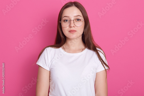 Close up portrait of beautiful young woman wearing spectacles and white casual outfit, feels happy, looks at camera, model posing isolated over pink studio wall, has long hair. People concept.