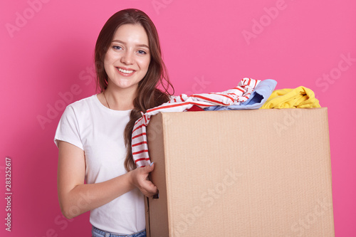 Quadro su tela Horizontal shot of female stands smiling near both with reusable clothing for poor peoplw isolated over pink background in studio, attractive woman making charity, posing with happy expression