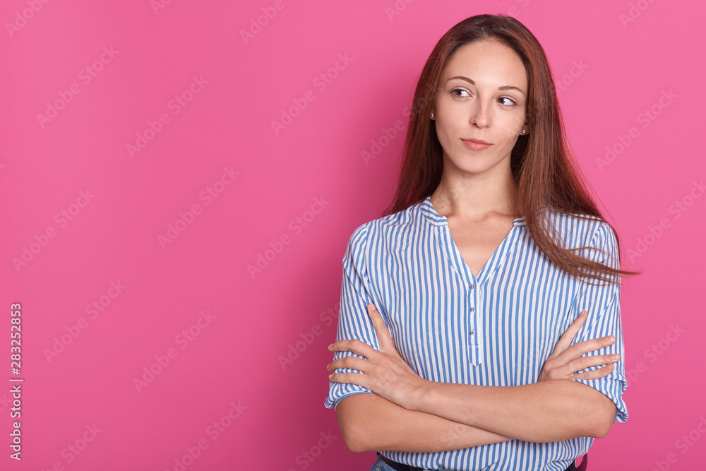 Portrait of young adorable woman in striped shirt looking aside, female holding hands crossed isolated over pink pastel wall background in studio. Copy space for advertismant or promotion text. Portrait of young adorable woman in striped shirt looking aside, female holding hands crossed isolated over pink pastel wall background in studio. Copy space for advertismant or promotion text.