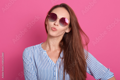 Wallpaper Mural Indoor shot of charming woman wearing stylish sunglasses and striped shirt, keeps lips rounded, making kiss gesture, model posing isolated over pink studio background. People emotions concept. Torontodigital.ca