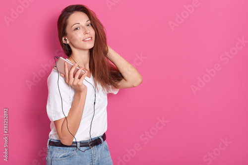 Cute dark haired girl listening to music via earphones on smartphone, having fun, wearing white t shirt and jeans, keeps hand brhind head, posing isolated over pink studio background. Copy space. photo
