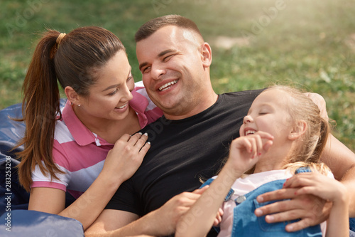 Young happy family spending free time in park on picnic, mother and father laugh from doughter's joke, they dressed casually, have happy facial expressions. Family, relationship and happyness concept. photo