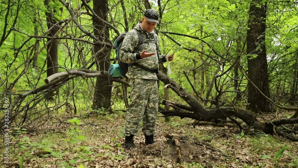 Caucasian handsome traveler in camouflage clothes, a cap and a backpack, walks through a green overgrown forest, and is guided by a map and a compass, medium plan Caucasian handsome traveler in camouflage clothes, a cap and a backpack, walks through a green overgrown forest, and is guided by a map and a compass, medium plan