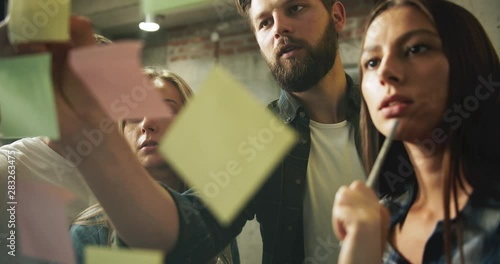 Team of modern creative office workers developing project plan and putting paper stickers on glass wall, advertisement campaign
