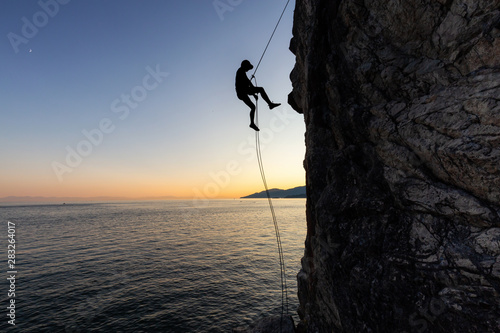 Silhouette of a Unrecognizable man rappelling down a steep cliff on the rocly ocean coast during a sunny summer sunset. Taken in Lighthouse Park, West Vancouver, British Columbia, Canada. photo