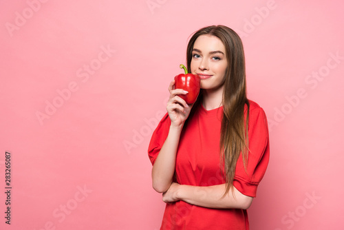 smiling attractive young woman holding red bell pepper and looking at camera on pink