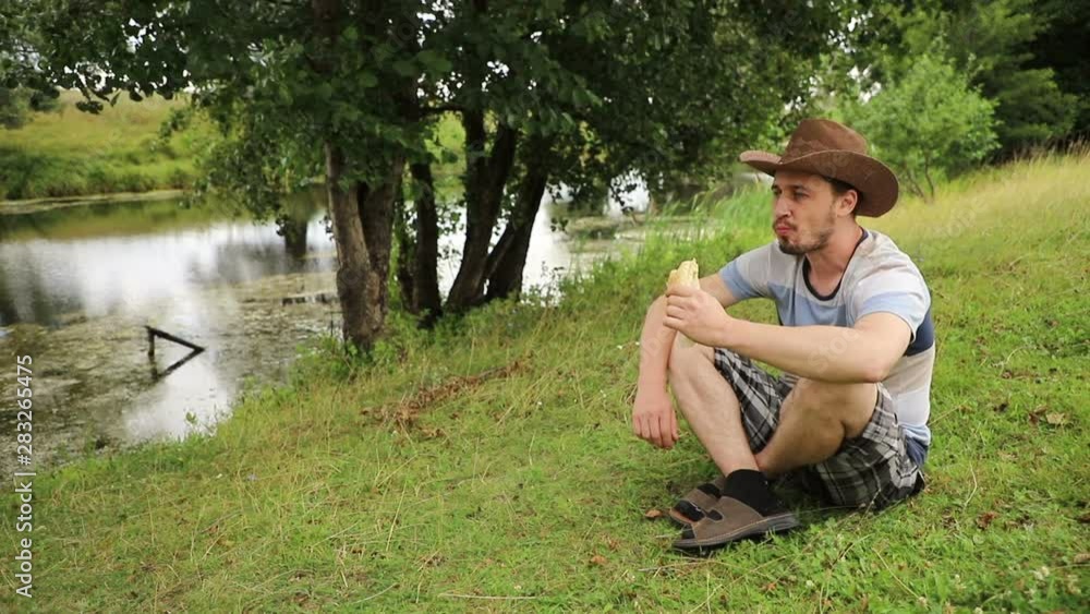 handsome caucasian man in a cowboy hat sits on a riverbank in cloudy weather and eats fast food, close-up handsome caucasian man in a cowboy hat sits on a riverbank in cloudy weather and eats fast food, close-up