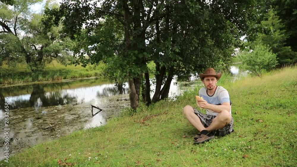 handsome caucasian man in a cowboy hat sits on a riverbank in cloudy weather and eats fast food, close-up handsome caucasian man in a cowboy hat sits on a riverbank in cloudy weather and eats fast food, close-up