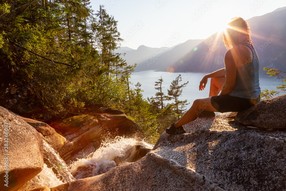 Adventurous Girl is sitting on top of a Beautiful Waterfall, Shannon Falls, and watching the sunset. Taken near Squamish, North of Vancouver, British Columbia, Canada. - obrazy, fototapety, plakaty 
