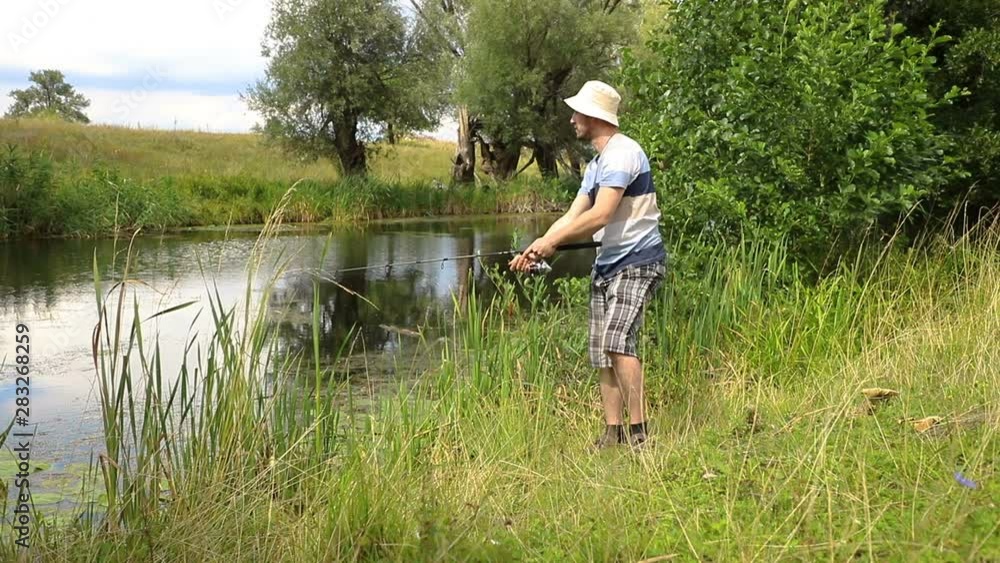 handsome a young caucasian fisherman in a panama hat throws a spinning rod into a river with green grass on the shore in cloudy weather, the average plan handsome a young caucasian fisherman in a panama hat throws a spinning rod into a river with green grass on the shore in cloudy weather, the average plan