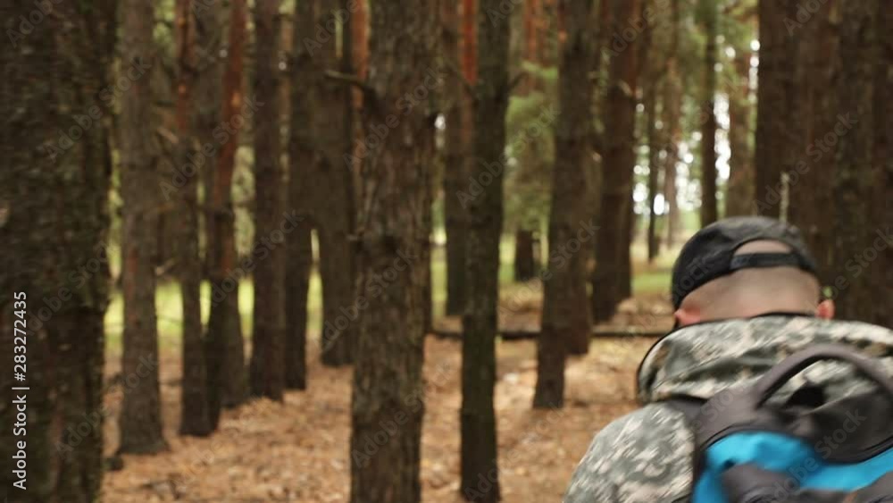 the camera moves behind the traveler in camouflage clothes, a cap and with a backpack, in a pine forest in clear weather, rear view, close-up the camera moves behind the traveler in camouflage clothes, a cap and with a backpack, in a pine forest in clear weather, rear view, close-up