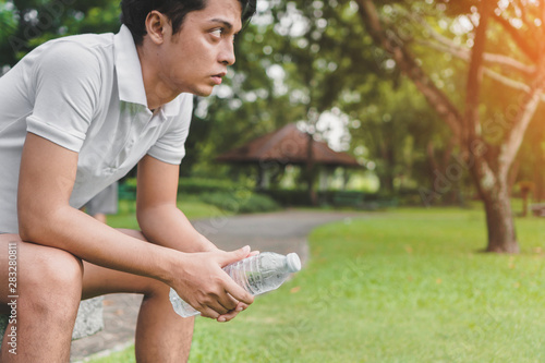 Young asian man runner relaxing holding drinking water bottle and sitting on bench in the park outdoors after sport at early morning time, Exercise and healthy concept.