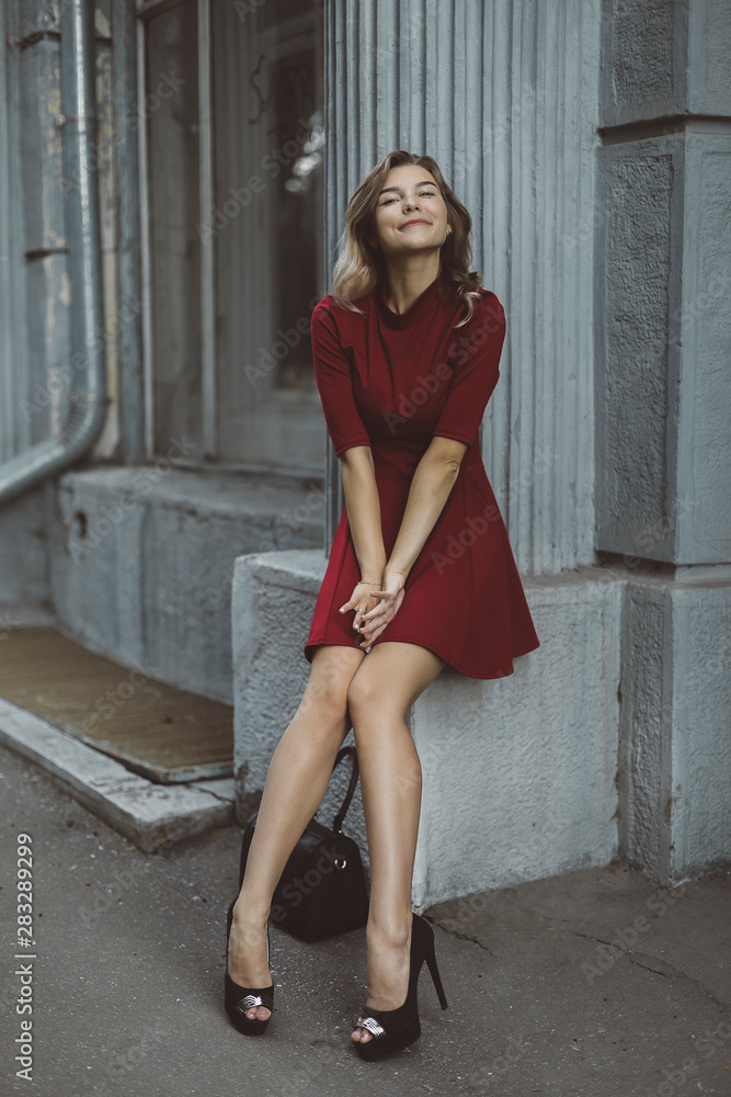 Naklejka premium Nice tanned stylish brunette girl in red short dress with flowers is standing poses and flirting walking on the street city