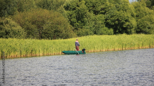 Summer water landscape, a lone fisherman on a small green boat with outboard motor stands with a fishing rod at the grassy river Bank