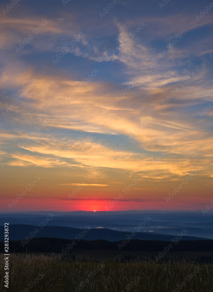 Sun at the moment of rising above the horizon with brightly colored sky slightly cloudy above hills, forests and meadows