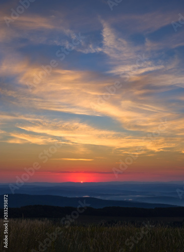 Sun at the moment of rising above the horizon with brightly colored sky slightly cloudy above hills, forests and meadows