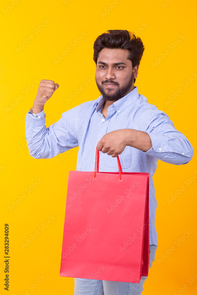 Young handsome Indian man with a beard is holding red shopping bags posing on a yellow background. Supermarket shopping concept. Young handsome Indian man with a beard is holding red shopping bags posing on a yellow background. Supermarket shopping concept.