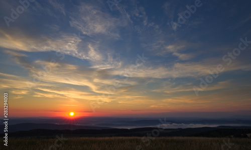 Sun right after rising above the horizon with brightly colored sky slightly cloudy above foggy hills © Martin