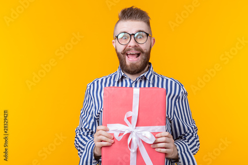 Joyful young hipster guy with a mustache in glasses hugs a red gift box on a yellow background. The concept of the joy of gifts. Advertising space.