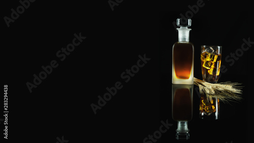 a glass of iced whisky and a bottle of whisky with wheat on reflected floor on dark background
