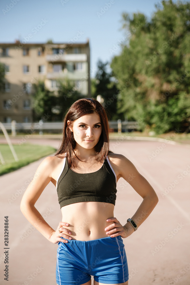 Portrait beautiful woman with a perfect figure in blue shorts and black top posing for the camera on running track at the stadium