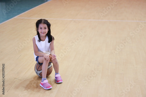 full shot of happy asian daughter taking a break and sitting on basketball in sport club with happy smiling face and looking at camera