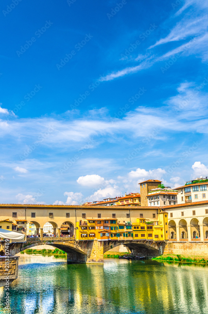 Ponte Vecchio stone bridge with colourful buildings houses over Arno River blue reflecting water in historical centre of Florence city, blue sky white clouds, vertical orientation, Tuscany, Italy