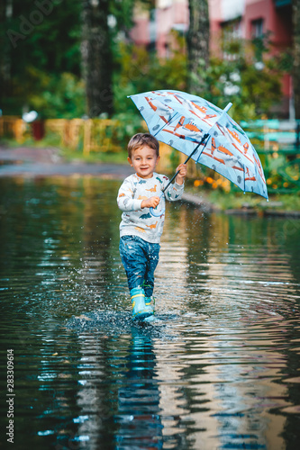 Happy little boy with colorful umbrella playing in puddle in rubber boots. A boy have fun after a rain.