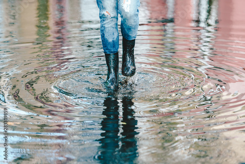 Womans feet with black rubber boots and blue jeans standing in a puddle of water after rain on a city street. Front view