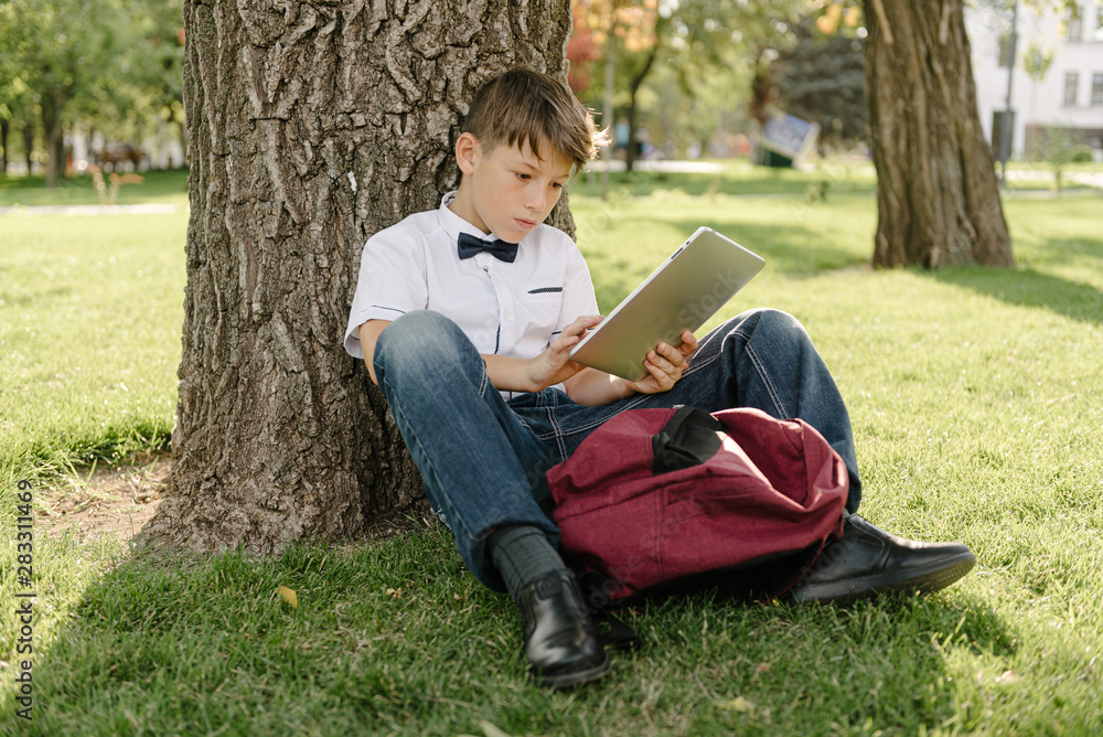 a schoolboy with a tablet sits on a lawn with his elbows on a tree. back to school a schoolboy with a tablet sits on a lawn with his elbows on a tree. back to school