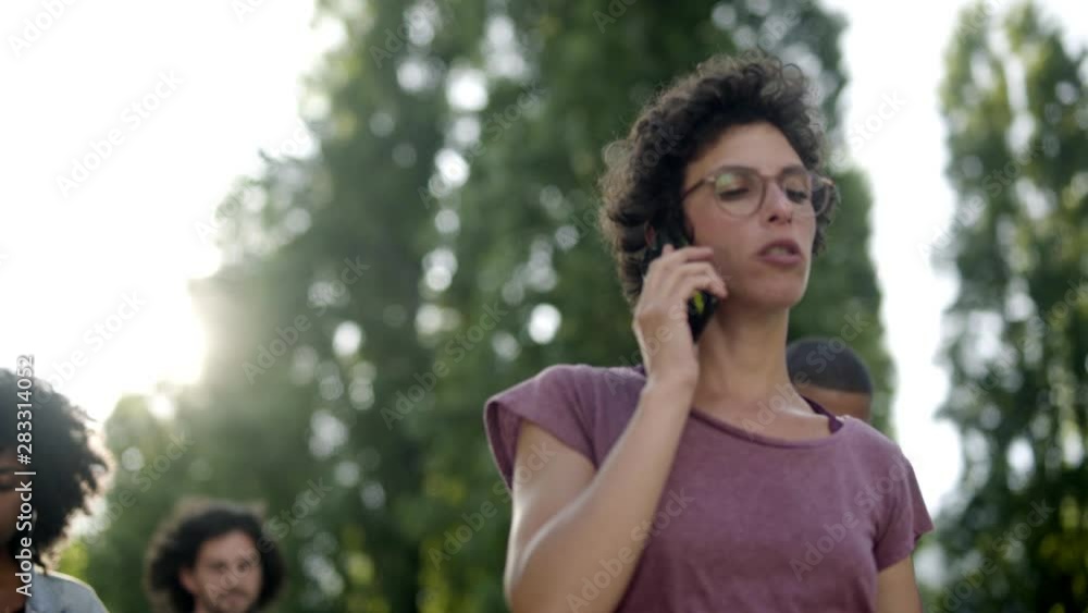 Serious young woman in eyeglasses talking on smartphone. Thoughtful curly lady with phone walking in park. Communication and technology concept Serious young woman in eyeglasses talking on smartphone. Thoughtful curly lady with phone walking in park. Communication and technology concept
