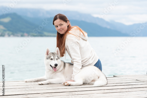 A young woman with brown hair and white sweater is sitting at the pier at the lake with calm water. A Siberian husky female dog is lying down near the girl. The Alps mountains in the background. © Rabinger