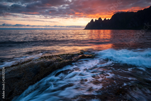 Waves crushing on dramtic rocks with Oksen mountains and colorful sunset in background, Tungeneset, Ersfjord, Senja, Norway