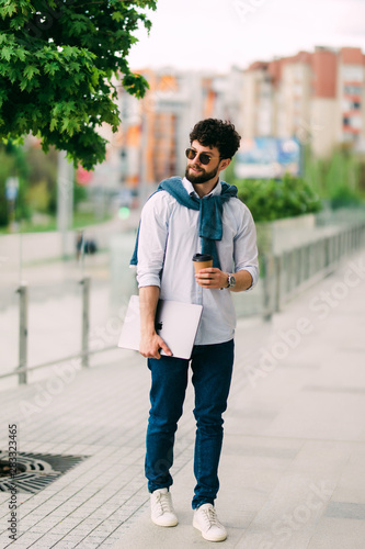 Hipster student going to university with laptop computer and cup of coffee. Bearded man wearing stylish casual clothes walking on the city street © F8  \ Suport Ukraine