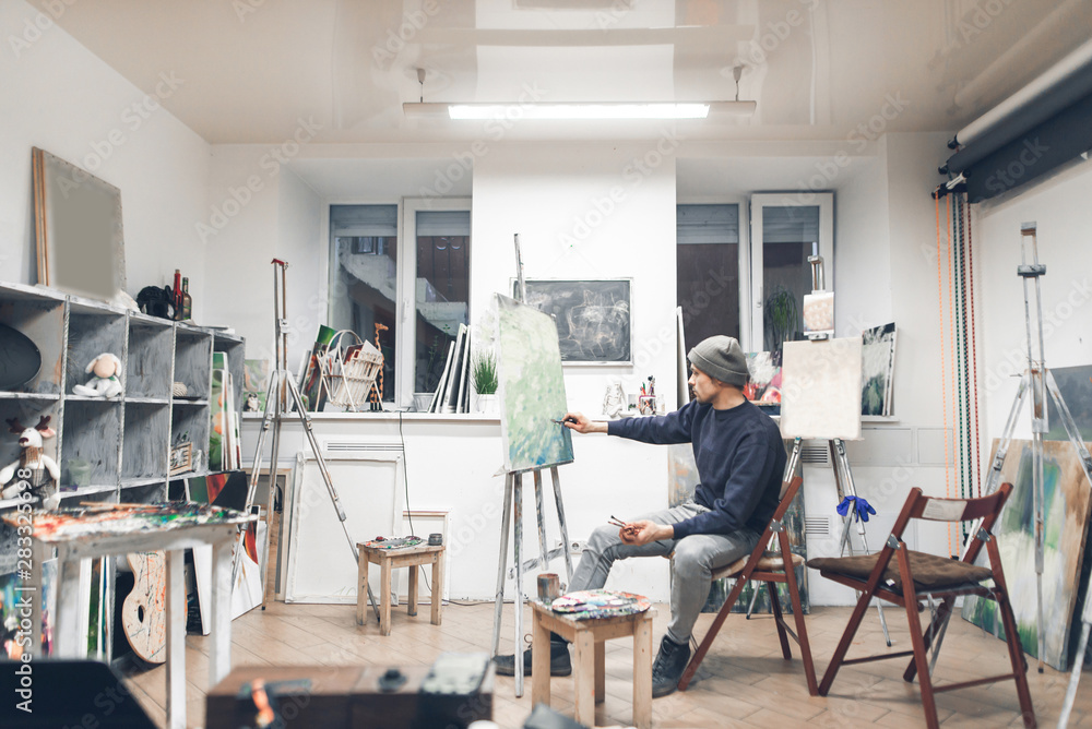 Professional artist sits on a chair in the interior of a cozy artistic studio and is working on a new painting. Painter paints a picture of oil in home studio. Atmosphere in the studio of painting.