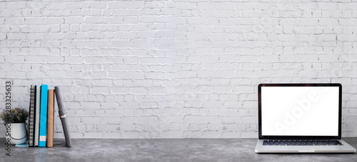 Blank screen on laptop with books and notebooks paper on brick wall texture background.