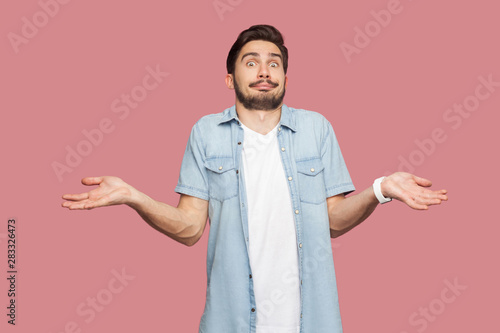I don't know. Portrait of confused handsome bearded young man in blue casual style shirt standing with raised arms and looking at camera with answer. indoor studio shot, isolated on pink background.