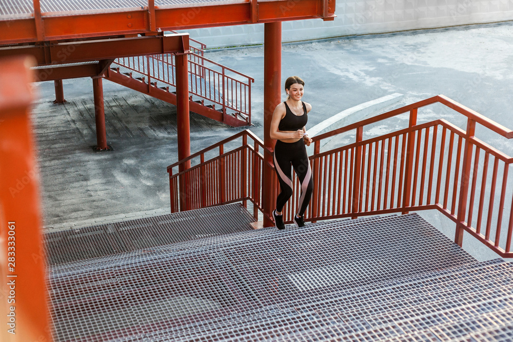 Young athletic slim beautiful woman running up stairs doing cardio interval training in black fashionable sportwear in the street on summer. Outdoor, sport and healthy concept