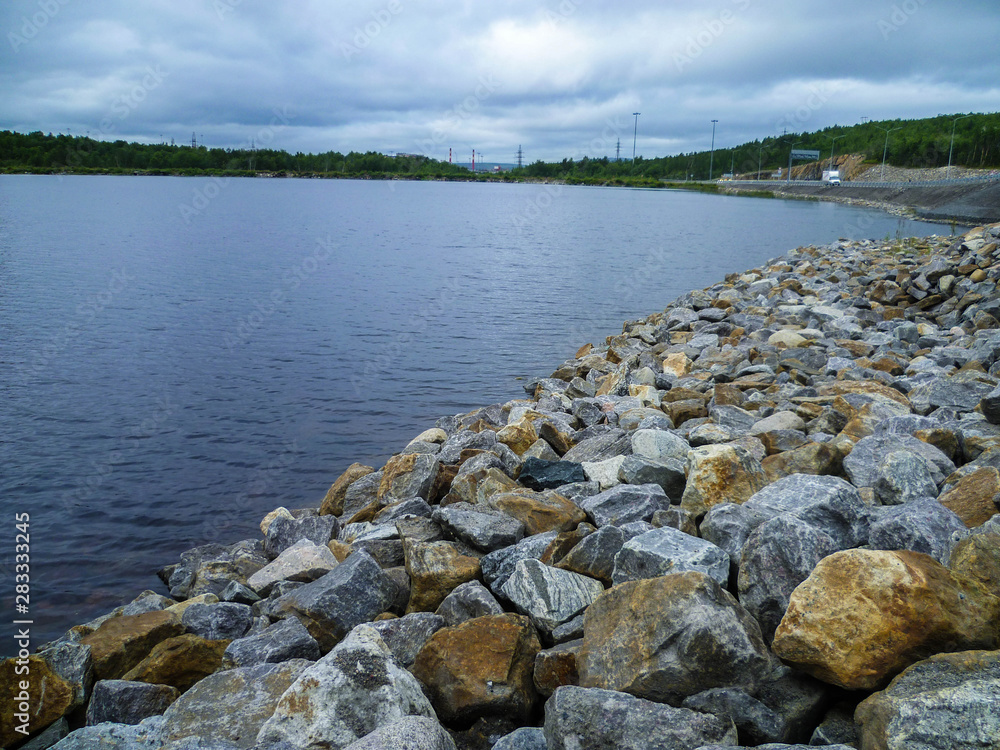 Fototapeta premium The shore of the lake, neatly stacked with large stones on a summer day.