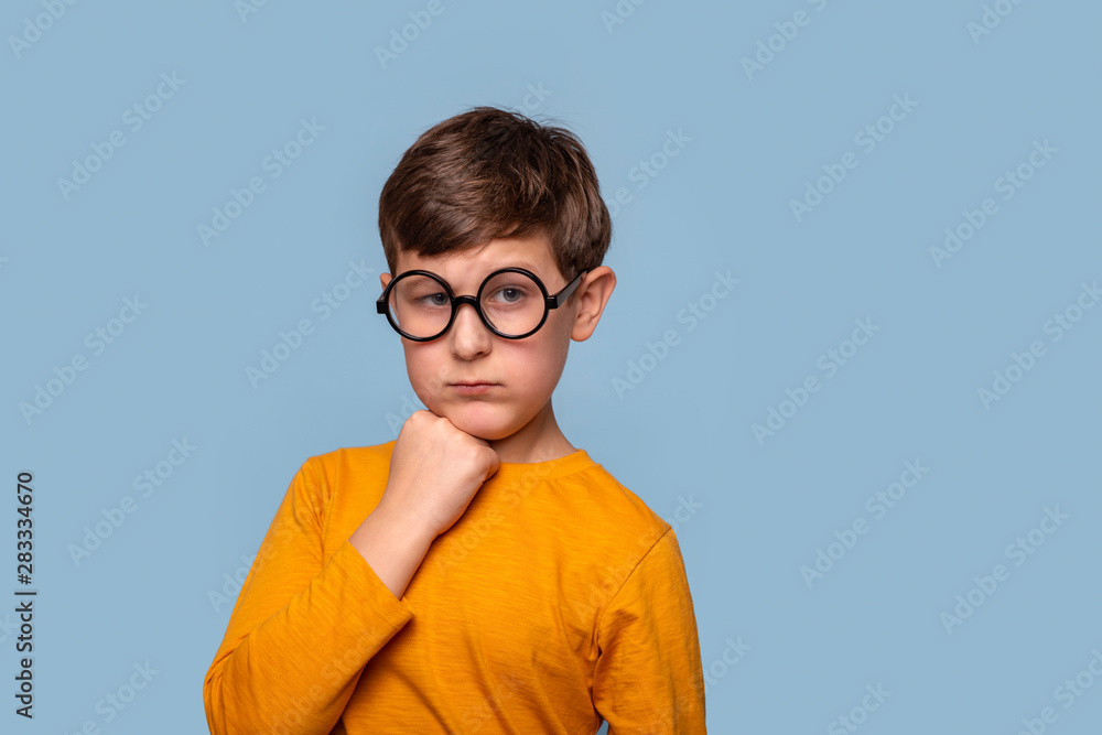 Waist up portrait of a young boy in round glasses thinking about something serious holding his chin with his fist , isolated on blue