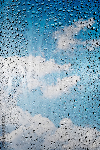 A blue sky with white cumulus clouds behind glass with flowing drops of water.