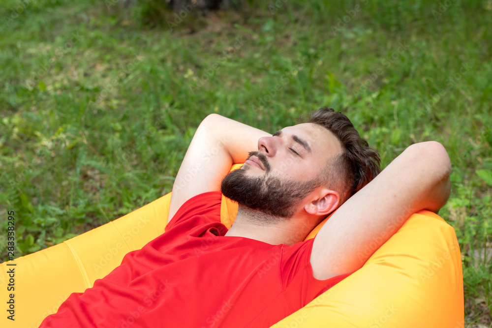 A young guy with a beard lies with his eyes closed on an orange inflatable sofa. The traveler is resting in nature.