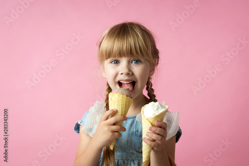 little girl with pigtails in a blue dress eating ice cream in a cone on a pink background